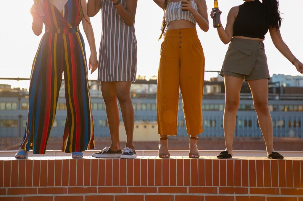 Group of crop unrecognizable multiracial young female friends in stylish clothes drinking bottles of beer while spending time together on rooftop during summer holidays